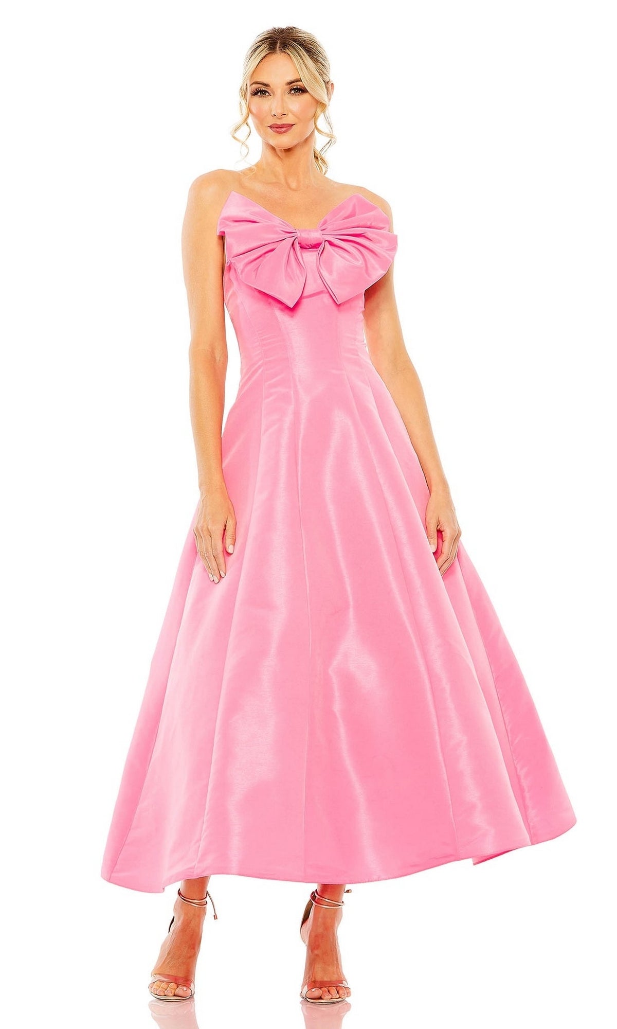 A woman stands against a plain white background in the Mac Duggal 49605, a tea-length, strapless pink wedding-guest dress with a large chest bow, paired with silver high-heeled sandals—a chic and elegant choice.